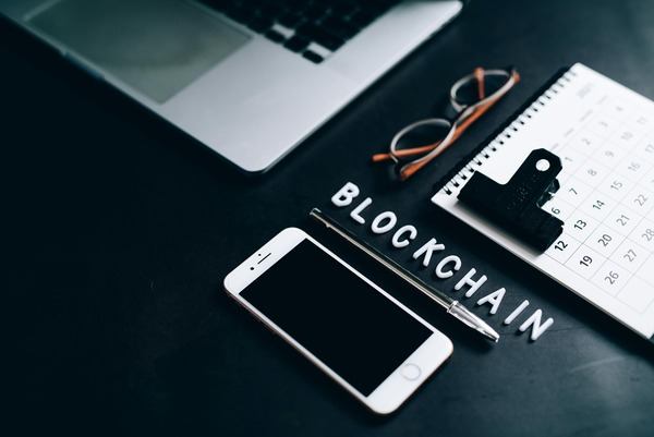 Notebook, phone and glasses next to the word blockchain on a dark desk